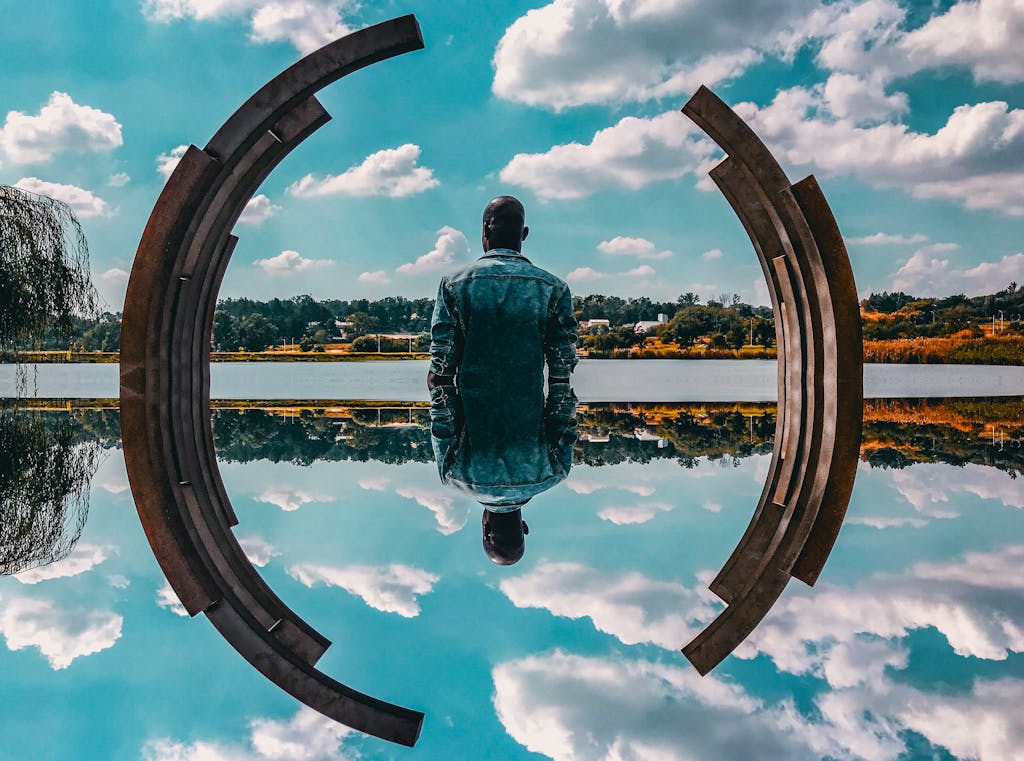A man in a denim jacket sits by abstract sculpture reflecting on the water under a blue sky.