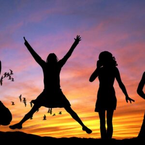 Silhouetted group of women joyfully jumping and posing against a vibrant sunset sky with birds flying.