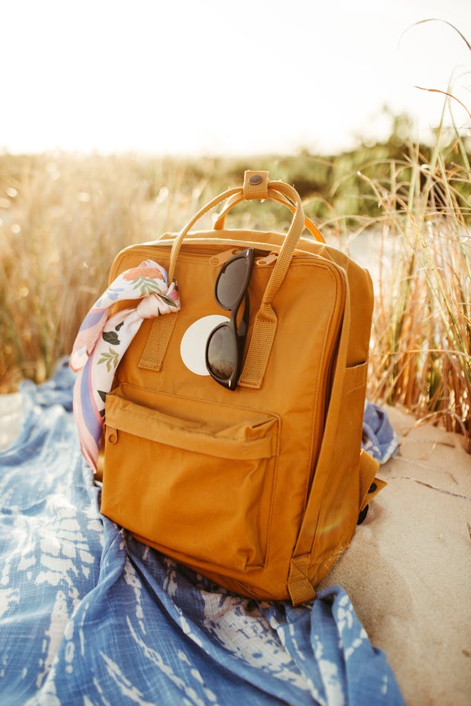 Stylish orange backpack with sunglasses resting on a beach blanket in summer sunlight.