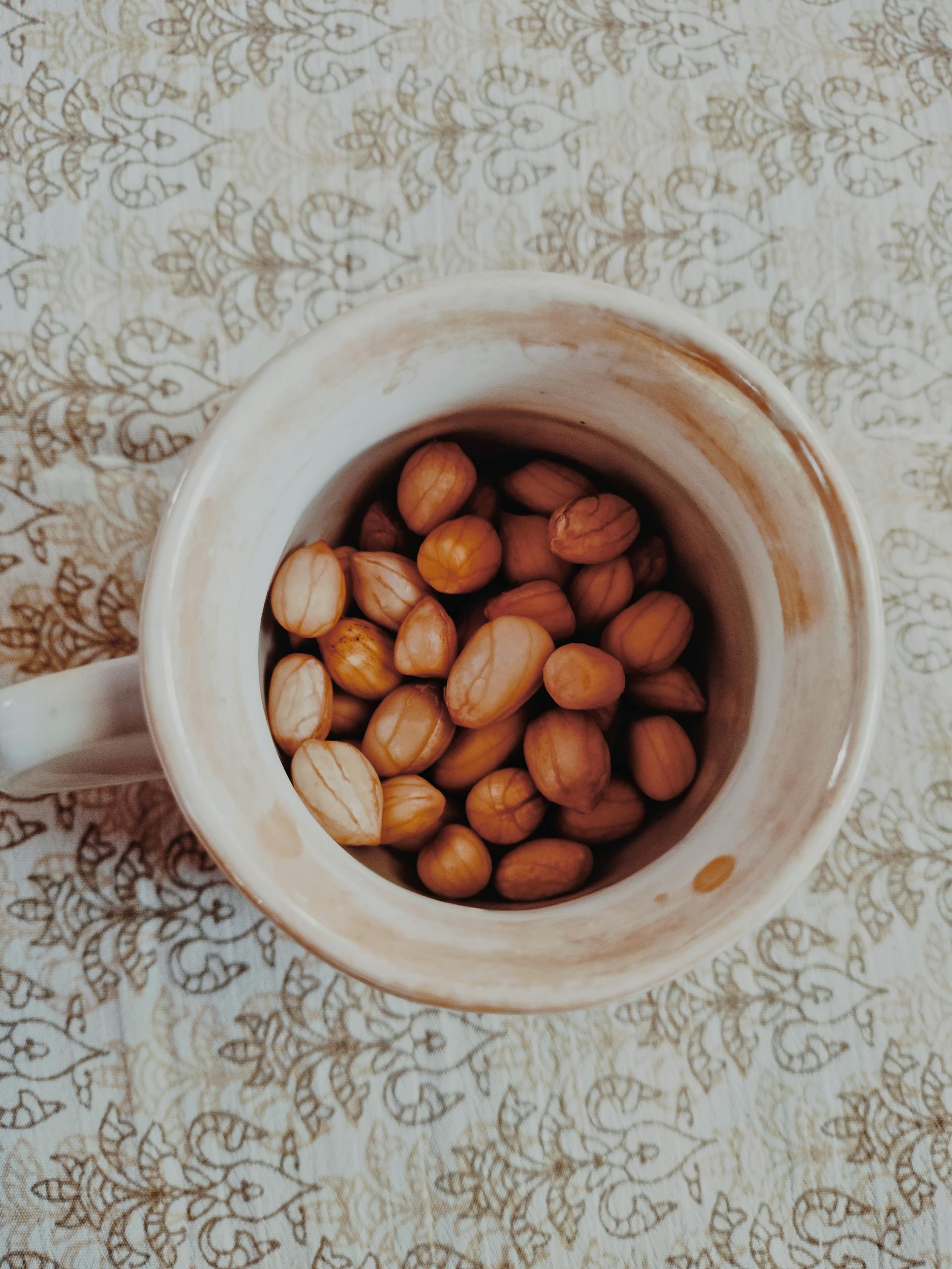 A ceramic mug filled with raw peanuts placed on a patterned tablecloth, top view.